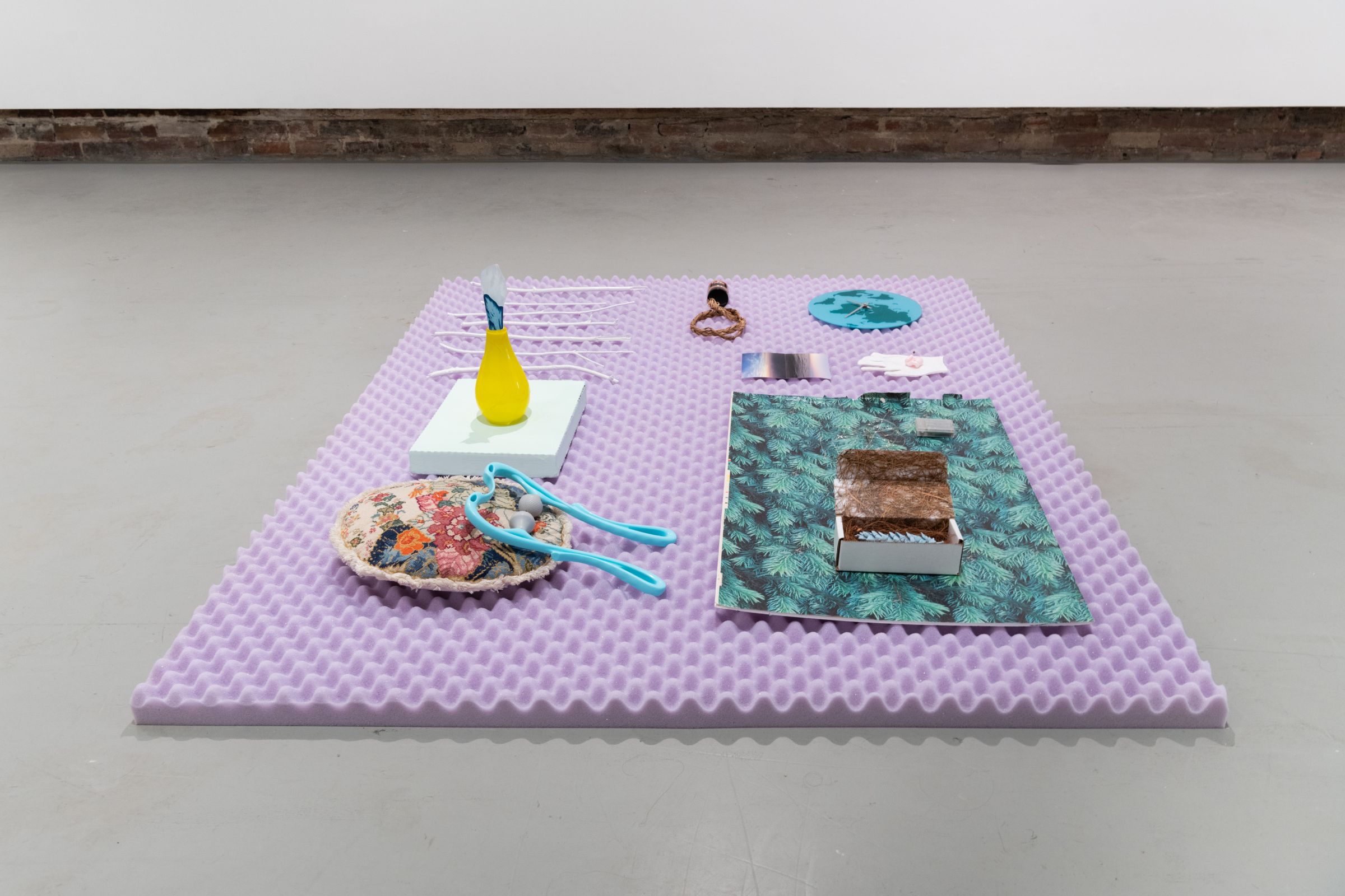 Floor sculpture made of purple mattress topper and various objects placed on top 