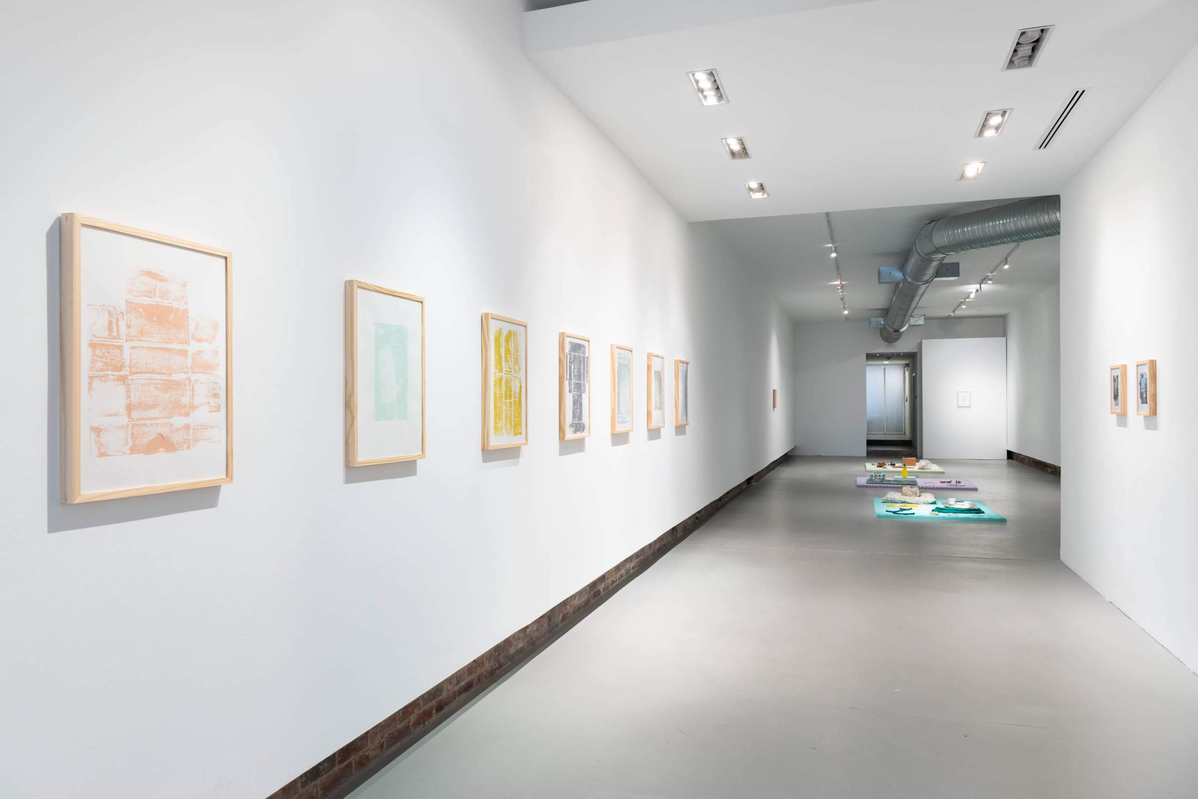 White-wall gallery space with framed prints on the left wall and sculptures on the floor in the background 
